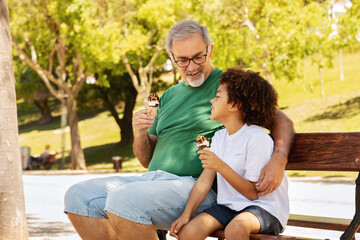 Wall Mural - Happy little mixed race boy and old caucasian grandfather enjoy free time, sit on bench, eating ice cream