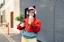 Hipster Fashion Woman In Bright Clothes, Heart Shaped Glasses, Headphones, Bucket Hat Drinking Fruity Flavored Tapioca Bubble Tea And Holding Green Potted Plant On The Gray Striped Wall Background
