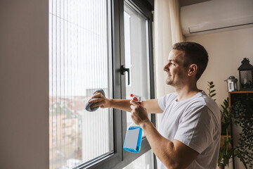 A tidy happy man is cleaning window glass at home with detergent and rug.