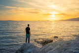 Fototapeta  - Young man standing on rocky beach at sunset time  