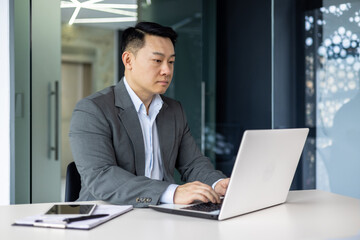Serious and thoughtful businessman working inside office sitting at table using laptop at work, mature asian boss in shirt thinking and typing on keyboard.
