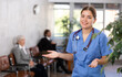 © JackF - Cheerful friendly young female doctor in blue uniform making welcome gesture, politely inviting patients in medical office