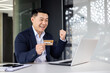 © Liubomir - Happy and successful businessman in office making online payments inside building, asian man using bank credit card and laptop for online shopping, man working sitting at desk