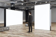 Businessman looks at blank white partitions with place for advertising poster or marketing campaign in stylish gallery hall with metallic benches on wooden floor and grey wall background, mockup