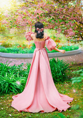 Wall Mural - Fantasy girl princess hand touching flowers sakura tree spring garden green grass petals fall. Woman queen back rear view long pink dress puffed sleeves satin bow in hair vintage old style art photo