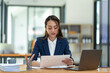 © crizzystudio - Asian businesswoman sitting happily in office holding detailed report paper analyzing data accuracy in financial matters management concept.