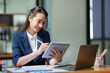 © crizzystudio - Asian businesswoman in a suit sitting holding iPad or tablet in contact See details and discuss business information happily in the office.