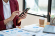 © crizzystudio - Beautiful smiling Asian businesswoman sitting analyzing charts and graphs showing changes in the market and holding smartphones at the office.