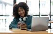 © crizzystudio - Young African businesswoman wearing headphones on her neck while looking at online shopping data Taking note of customer orders on a laptop while holding a mug of warm coffee to relax.