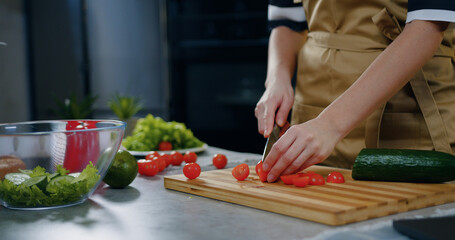 Unknown woman holding a knife in her hands and cutting tomatoes while cooking vegetable salad near kitchen table ,close up