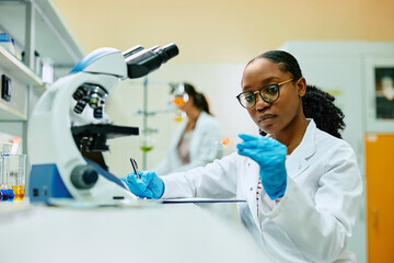 Wall Mural - young black biochemist examining sample on microscope slide during her research in lab.