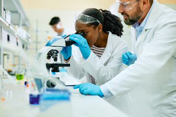 Young black scientist analyzing medical sample under the microscope while working with her colleague in lab.