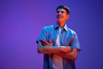 Portrait of happy, smiling, young guy in casual clothes posing with positive mood against gradient purple background in neon light. Delightful. Concept of human emotions, lifestyle, youth