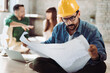 © Goran - Young architect working on blueprints in the office. There are people in the background