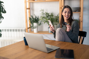 Happy young businesswoman, satisfied buyer holding credit card, using online bank app on smart mobile phone making convenient financial e-commerce payment digital transaction while sitting at home.