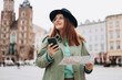 Beautiful stylish woman in hat walking on Market Square in Krakow on autumn day and holding mobile phone and map. Phone Communication. Urban lifestyle concept. Check social networks, booking hotel