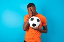 Man Wearing Orange T-shirt Holding A Ball Over Blue Background Covering Her Face With Her Hands, Being Devastated And Crying. Sad Concept
