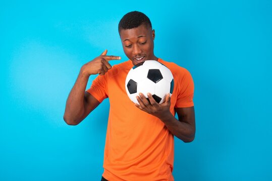 Shocked Man wearing orange T-shirt holding a ball over blue background look surprisedly down, indicates at blank  with fore finger, Scared model