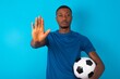 © Roquillo - Young man wearing sport T-shirt holding a ball over blue background shows stop sign prohibition symbol keeps palm forward to camera with strict expression