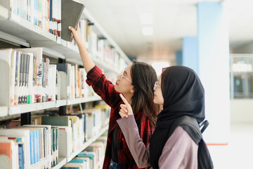 Two joyful smiling young student girls choosing books from the bookshelf in the school library. 