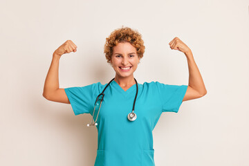 Happy curly young lady in turquoise medical uniform with stethoscope on her neck stands on white background, clenching her fists up with joy, good doctor concept, copy space