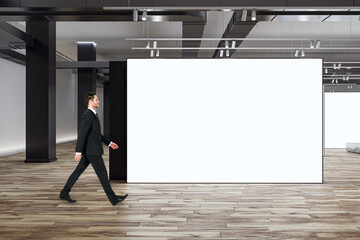 Marketing concept with businessman walking by blank white partition with space for advertising poster or picture frame in loft style gallery hall with wooden floor and grey wall background, mock up