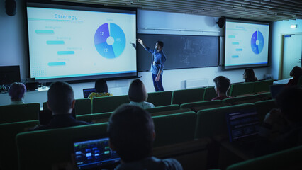 Fototapete - Young Male Teacher Giving a Business Strategy Lecture to Diverse Multiethnic Group of Female and Male Students in Dark College Room. Projecting Slideshow with Comparison Charts and Commercial Data