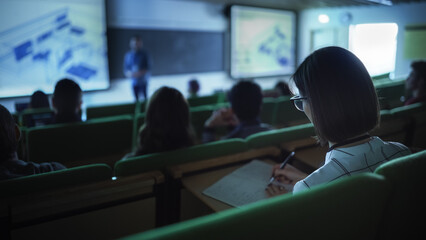 Fototapete - Over the Shoulder Footage of a Female Student Taking Down Notes in a Notebook, Sitting in College Auditorium on a Lecture. Young Woman with Short Hair Wearing Glasses and Studying in University