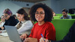 © Gorodenkoff - Portrait of an Empowered African Female Student Studying in University with Diverse Multiethnic Classmates. Young Happy Black Woman Looking at Camera and Smiling. Using Laptop Computer in Class