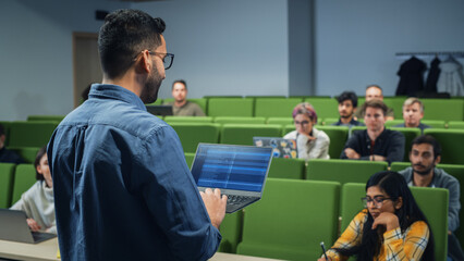 Fototapete - University Professor Explaining the Importance of a College Degree to a Group of Diverse Students in a Modern Auditorium. Teacher Holding a Laptop Computer and Talking to Scholars. Camera Facing Class