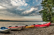 North Hatley, QC, Canada - September 5, 2014: Red, yellow, blue and green kayaks sit on a pebbly beach beside a stand of trees as the sun rises with dramatic clouds over Lake Massawippi