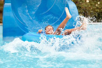 Wall Mural - Joyful boy in swimming goggles slides down the blue water slide on an inflatable boat in the water park, water splashes, joyful mood, summer vacation