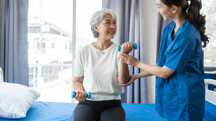 Young physiotherapist helping senior mature asian woman grey hair work out with dumbbells, to recover from injury at health centre in physical therapy session.