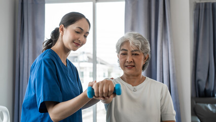 Young physiotherapist helping senior mature asian woman grey hair work out with dumbbells, to recover from injury at health centre in physical therapy session.