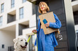 Young woman picks up parcels from automatic post office machine, standing with her dog near apartment building. Concept of fast delivery and urban lifestyle