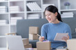 © Tj - Asian business woman siting on sofa using a laptop and notebook checking order online shipping boxes at home. Starting SME Small business entrepreneur freelance. Online business, SME Work