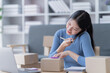 © Tj - Asian business woman siting on sofa using a laptop and notebook checking order online shipping boxes at home. Starting SME Small business entrepreneur freelance. Online business, SME Work