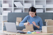© Tj - Asian business woman siting on sofa using a laptop and notebook checking order online shipping boxes at home. Starting SME Small business entrepreneur freelance. Online business, SME Work