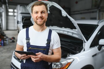 Bearded mechanic in overalls standing in garage of a car salon and holding tablet. He is about to diagnostic breakdown.