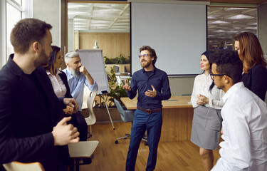 Young man business coach trainer talking to group of people in office standing around him and listening. Office workers, entrepreneurs on corporative training, team building seminar or master class.