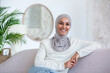 © Liubomir - Portrait of young business woman of oriental origin in hijab sitting on sofa at home and looking at camera confidently and smiling.