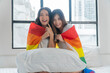 © kaew6566 - Romantic asian couple woman hugging each other together with rainbow flag sitting bed beside window. Cheerful lover lesbian casual girl with lgbt flag hug closeness at home. LGBTQ couple relationship