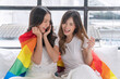 © kaew6566 - Romantic asian couple woman hugging each other together with rainbow flag sitting bed beside window. Cheerful lover lesbian casual girl with lgbt flag hug closeness at home. LGBTQ couple relationship