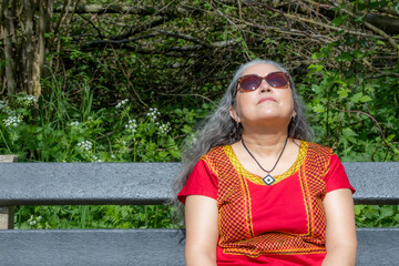 Wall Mural - Brunette woman sitting with eyes closed sunbathing on wooden bench, wild green vegetation in blurred background, native yellow red blouse, sunglasses, gray wavy gray hair, enjoying sunny day