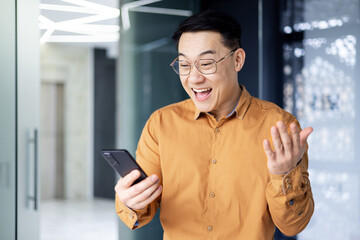 Wall Mural - Asian man using phone standing near window inside office building, programmer in shirt and glasses celebrating victory and successfully completed software development, reading news from smartphone.