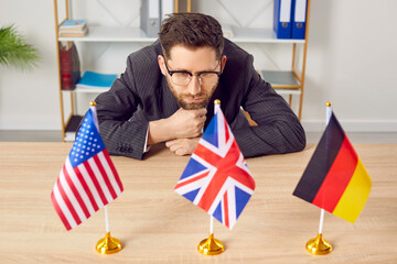 Pensive businessman sitting at desk looking at flags of different countries. Young man in formal wear choosing foreign language or country to work or study in. USA, Great Britain and Germany flags