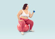 Fat woman doing fitness workout with sports equipment. Plump chubby woman in bra top and pink leggings sitting on red fitball and training muscles with dumbbell isolated over light blue background