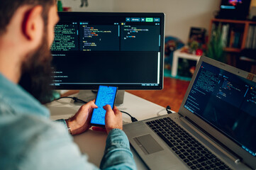 View from behind of an operating data security worker reading text on his phone while sitting at home office surrounded by monitors.