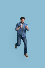 Handsome smiling man holding mobile phone shopping online jumping high isolated on blue background. Happy hipster wearing stylish casual clothes, shoes running fast, hurry up