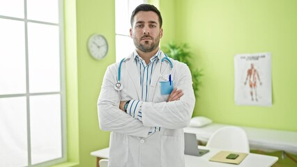 Poster - Young hispanic man doctor standing with serious expression and arms crossed gesture at clinic